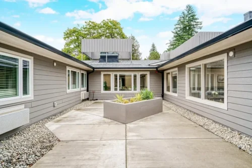 Contemporary courtyard with gray siding, large windows, concrete path, and a central planter surrounded by greenery under a blue sky.