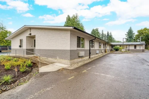 A modern single-story building with gray brick exterior, surrounded by greenery, and a clear blue sky.