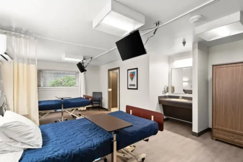 A hospital room with two hospital beds covered in blue blankets and adjustable overbed tables. A wall-mounted TV, a chair, and a sink are visible, conveying a clean and functional atmosphere.