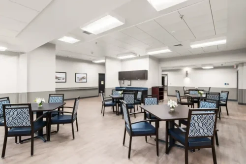 A modern dining area with patterned blue chairs around square dark tables. Soft lighting, neutral walls, and floral centerpieces create a calm atmosphere.