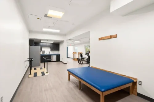 Bright physical therapy room with a blue treatment table, parallel bars, and exercise equipment. Modern kitchen area in the background. Clean, clinical feel.